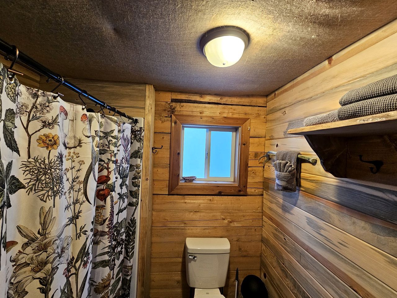 A small, wooden cabin bathroom with a toilet, a window, and a shower curtain with a floral pattern.
