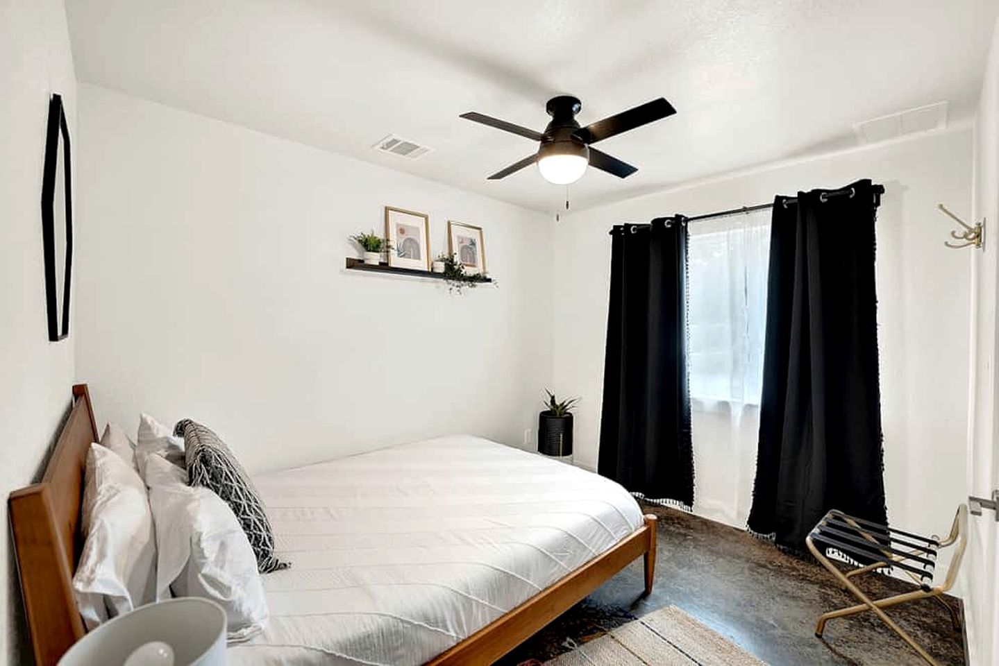 Un dormitorio con cama, ventilador de techo, ventana con cortinas negras, estante pequeño con fotografías y una silla.