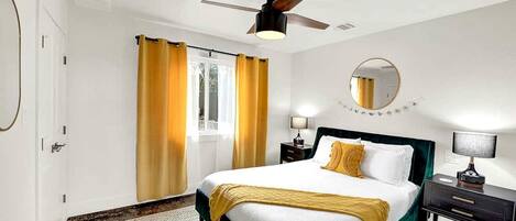 A hotel room with a bed, a ceiling fan, a window with curtains, a bedside table with a lamp, and a round mirror on the wall.