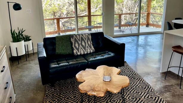 A living room with a black leather sofa, a wooden coffee table, and a patterned rug. There are large windows with a view of trees and a balcony. - Luxury Cabin for Four for Texas Glamping Retreat (Wimberley)