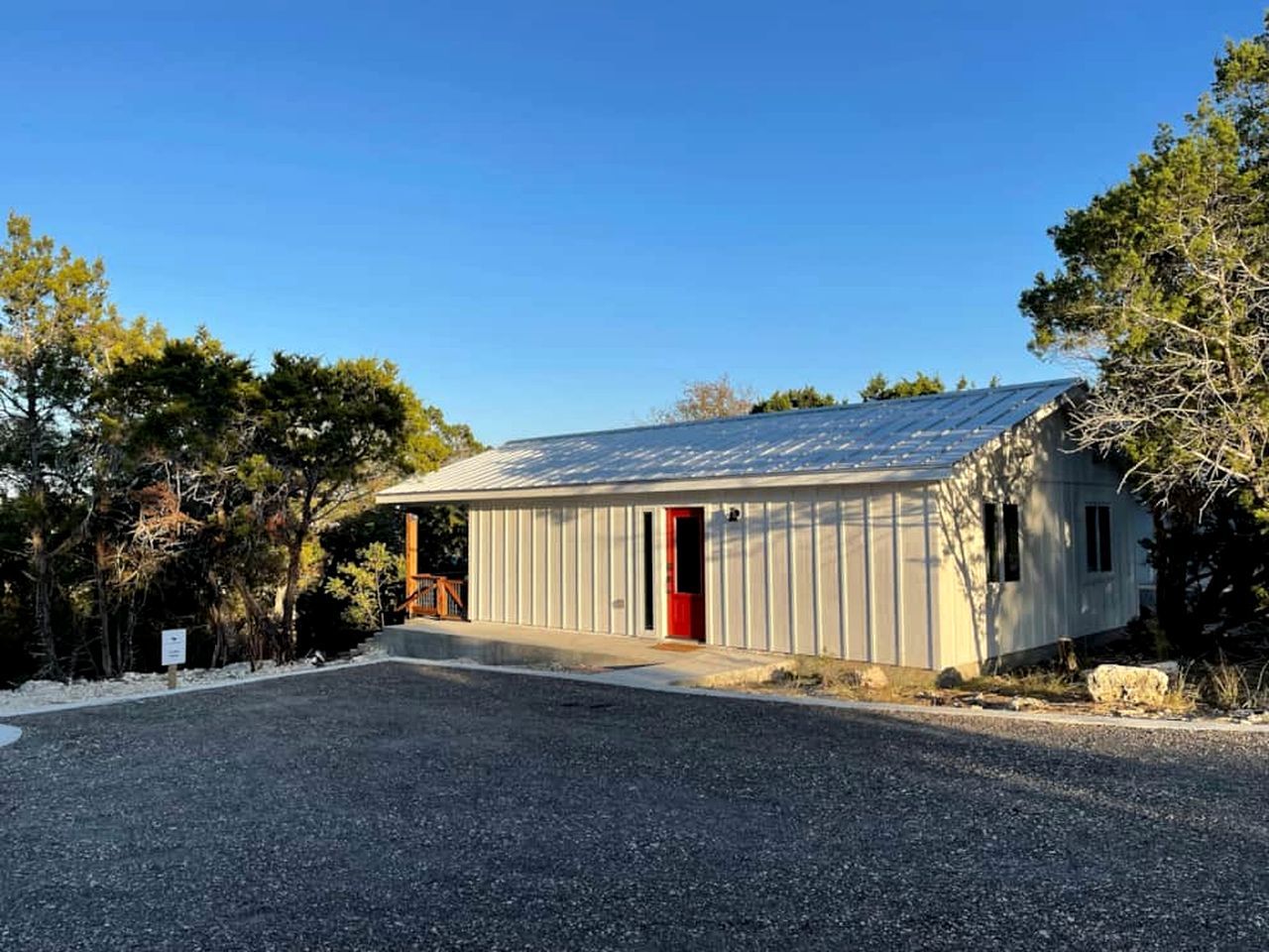 A single-story building with a red door, a gravel driveway, and surrounding trees.