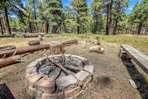 A campsite with a fire pit surrounded by logs and rocks, a wooden bench, and a forested background. - Gorgeous Historic Cabin with Grill and Fire Pit Surrounded by Pines for a Lovely Outdoor Experience in Parks, Arizona (Parks)