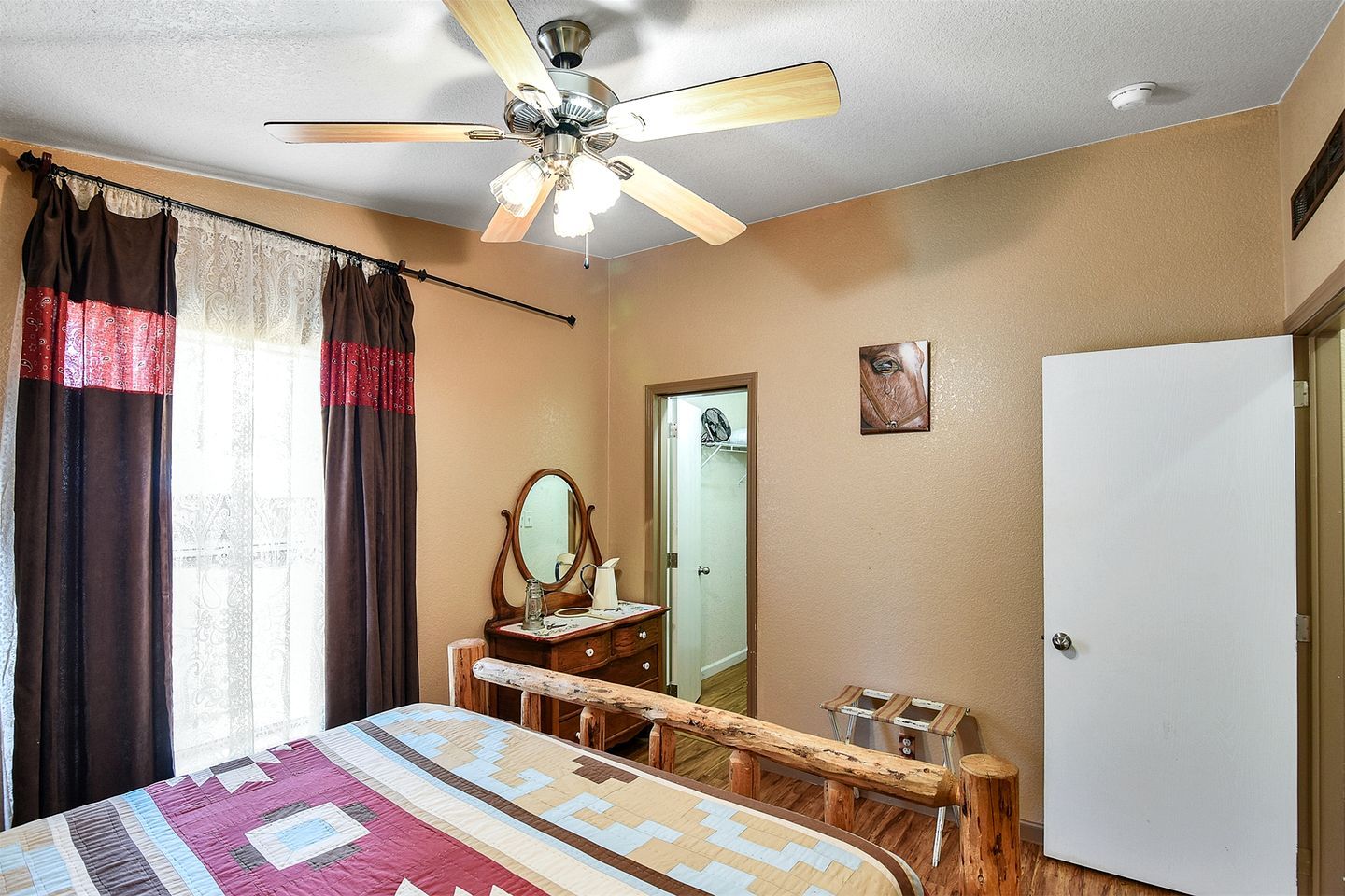 A bedroom with a bed, a wooden bench, a ceiling fan, a mirror, a door, and a white wall.