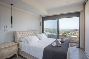 A bedroom with a large bed, a nightstand, a dresser, and a view of the city through a large window. - Aetheria Luxury Villa (Καλυθιές)