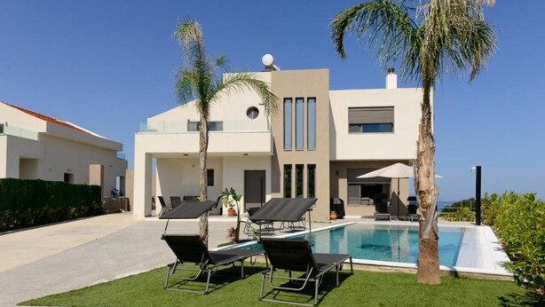 Uma casa moderna de dois andares com piscina, duas espreguiçadeiras e palmeiras.