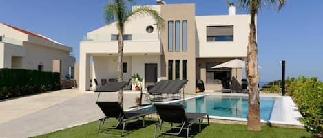 A modern two-story house with a swimming pool, two lounge chairs, and palm trees.