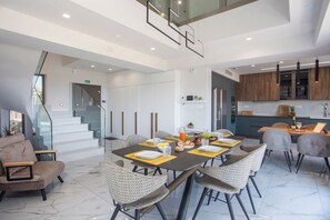 A modern dining area with a high ceiling, a staircase, and a kitchen with wooden cabinets. - Aetheria Luxury Villa (Καλυθιές)