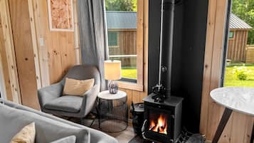 A cozy room with a wood-burning stove, a grey armchair, a small round table with a lamp, and a view of the outdoors.