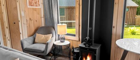 Una habitación acogedora con estufa de leña, un sillón gris, una mesita redonda con lámpara y vistas al exterior.