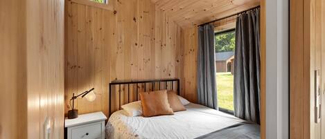 Un dormitorio con paneles de madera, una cama, una mesita de noche, una lámpara y vistas al exterior.