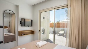 A hotel room with a bed, a TV mounted on the wall, a mirror, and a sliding glass door leading to an outdoor area with a chair and umbrella. - Aura Luxury Villas Anasa, Villa (Rhodes)