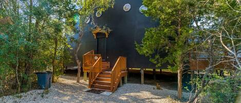 A house with a wooden staircase leading to a door, surrounded by trees and gravel.