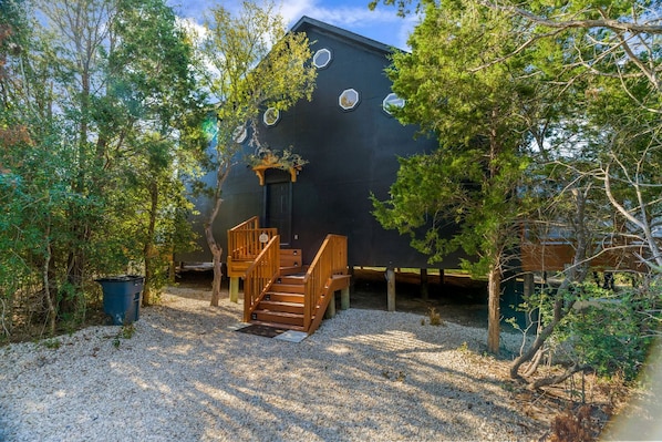 A house with a wooden staircase leading to a door, surrounded by trees and gravel.