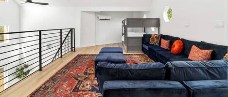 A modern living room with a bunk bed, a blue sofa set, a patterned rug, and a balcony with a railing.