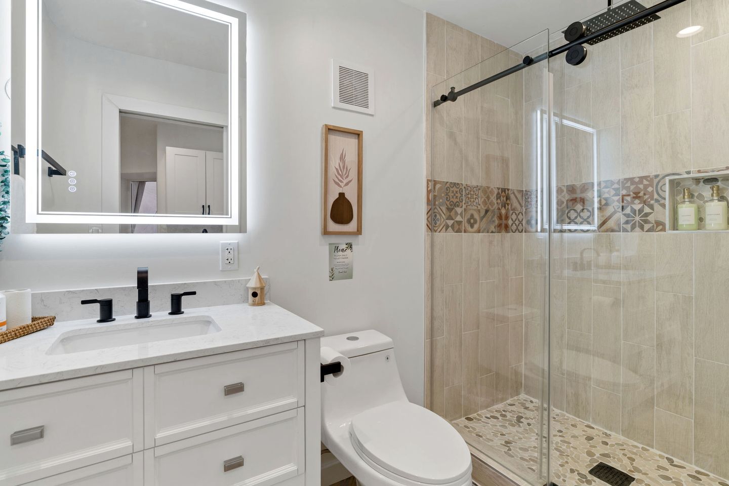 Een badkamer met een wit wastafel, een zwarte kraan, een toilet, een douche met een glazen deur, en een spiegel met verlichting.
