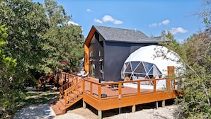 Una cabaña moderna con una terraza de madera, una gran estructura en forma de cúpula y una escalera que lleva a la entrada.