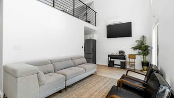 Una sala moderna con un sofá seccional gris, un escritorio pequeño negro, un televisor montado en la pared, una escalera y una planta en maceta.