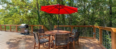 Una terraza de madera con una mesa redonda, sillas, un paraguas rojo y una parrilla.