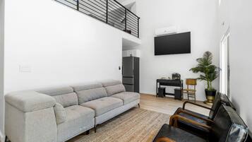 Een moderne woonkamer met een grijze hoekbank, een klein zwart bureau, een aan de muur bevestigde televisie, een trap en een potplant.