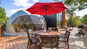 Une structure en forme de dôme géodésique dotée d’une terrasse en bois, de sièges extérieurs et d’un parasol rouge.
