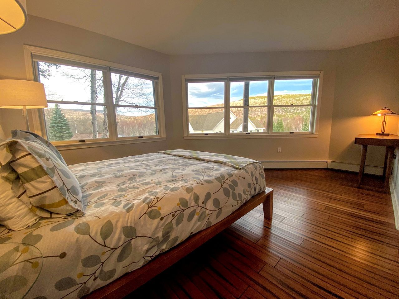 A bedroom with a bed, two windows, a lamp, and a wooden floor.