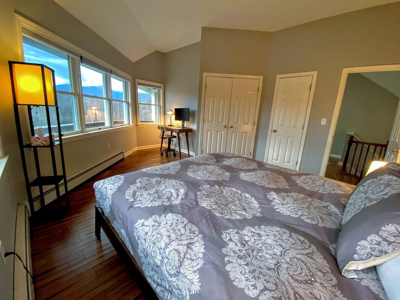 A bedroom with a large bed, a desk with a chair, a standing lamp, and a view of the outdoors through the windows.