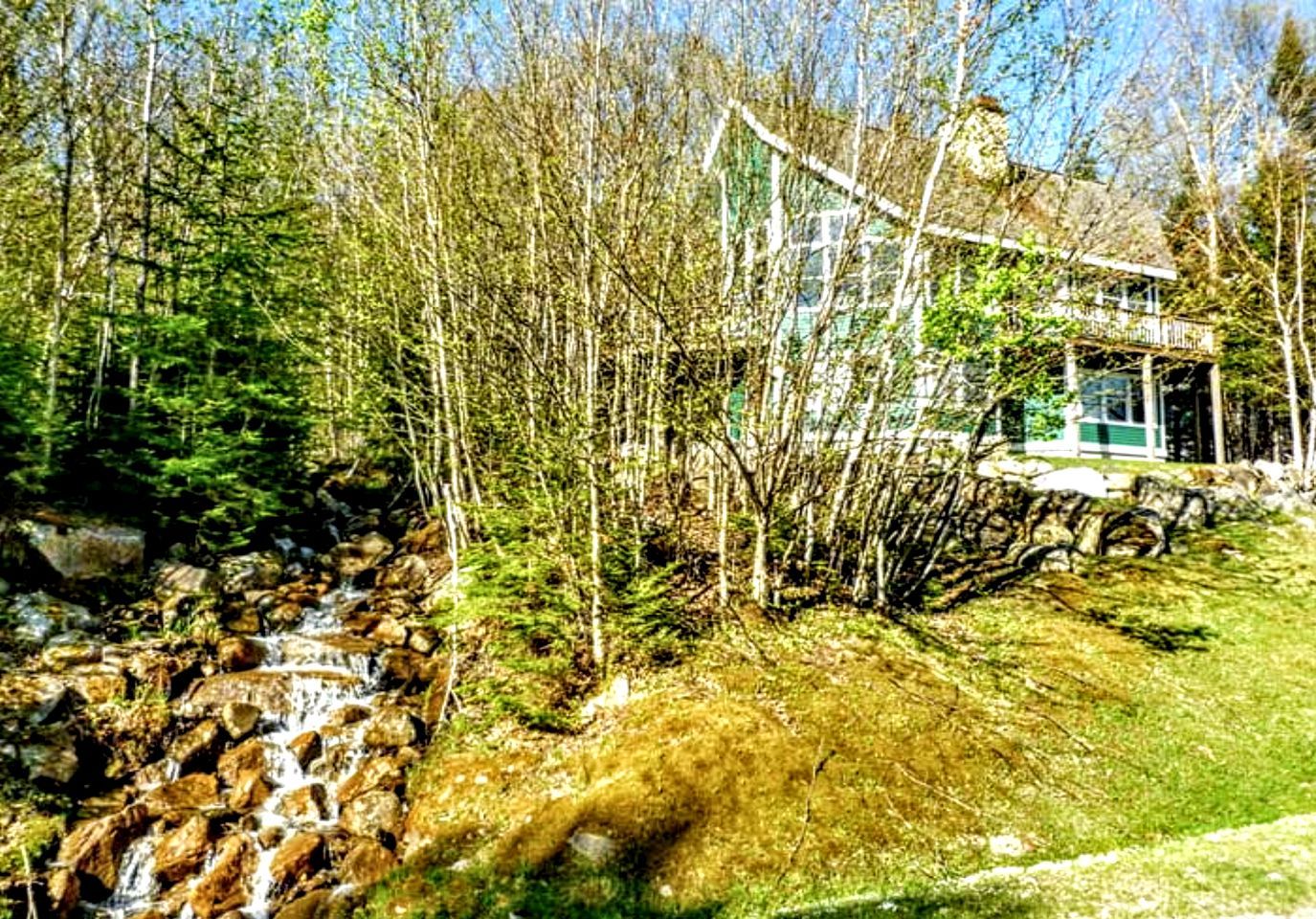 A house with a stone pathway leading to a small waterfall, surrounded by dense trees and a clear sky.