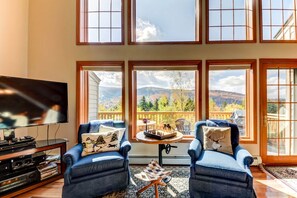 A living room with a TV, two blue armchairs, a coffee table, and a view of mountains through large windows. - Bretton Woods Vacation Rental near Mt Washington (Carroll)