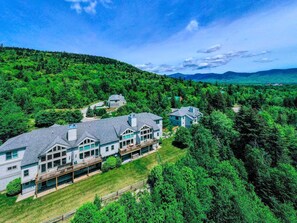 A large, multi-story house with a spacious deck, surrounded by dense greenery and hills in the background. - Bretton Woods Vacation Rental near Mt Washington (Carroll)