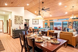 A dining area with a wooden table set for a meal, a pool table, and a sofa with a view of the outdoors. - Experience Bliss: Big Bear Lake Rental with Exclusive Hot Tub (Big Bear Lake)