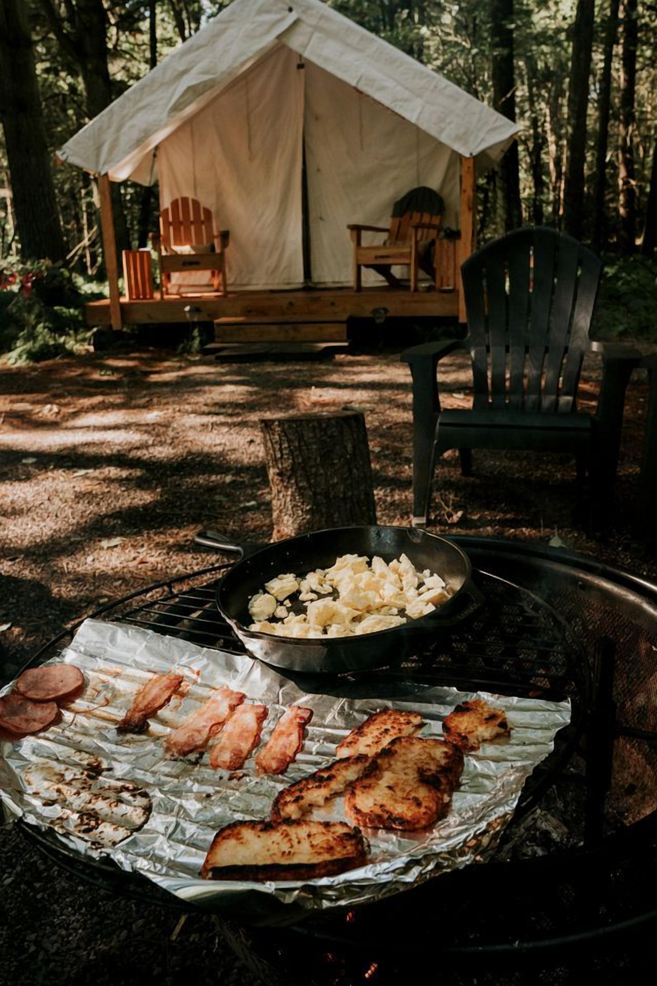 Ein Zelt mit Esstisch und Stühlen, ein Grill mit Essen und eine bewaldete Fläche.