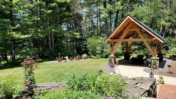 A wooden gazebo with a sloped roof, surrounded by a garden with various plants and a lawn.