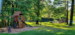 Un parque infantil de madera con columpios y un tobogán, rodeado de árboles y una zona de picnic.