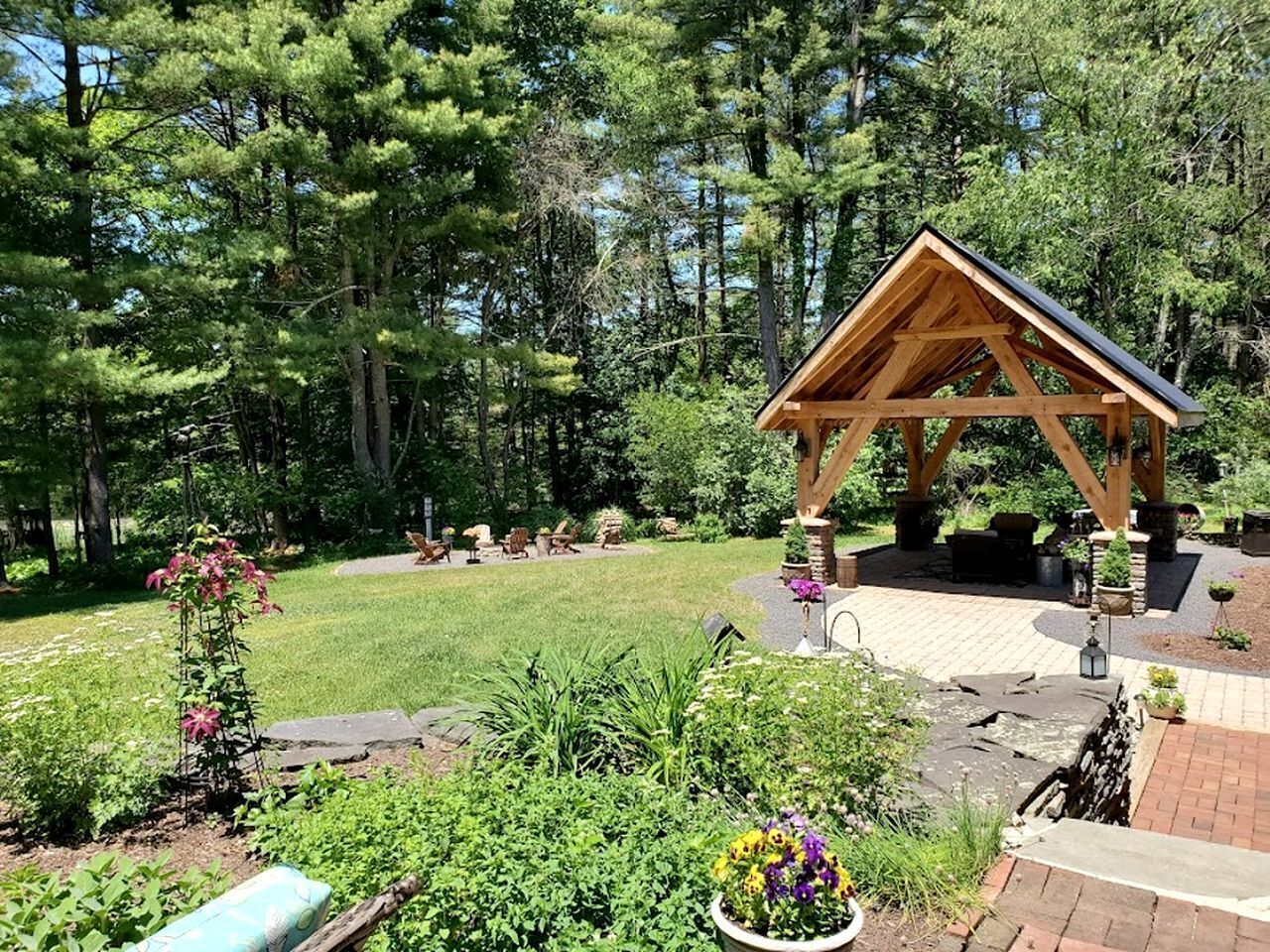 A wooden gazebo with a sloped roof, surrounded by a garden with various plants and a lawn.
