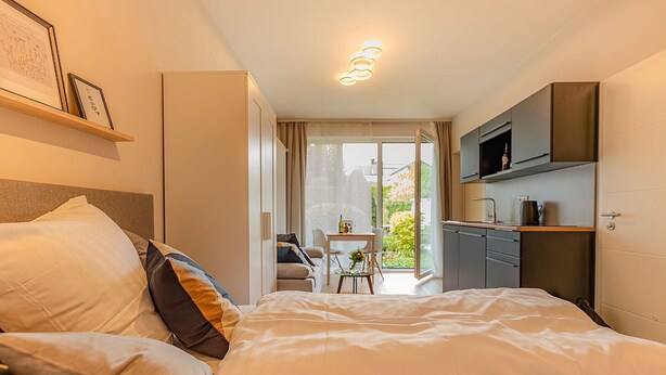 A modern hotel room with a bed, a small table with chairs, a kitchenette, and a balcony with a view. - Studio at the Vineyards (Iphofen)