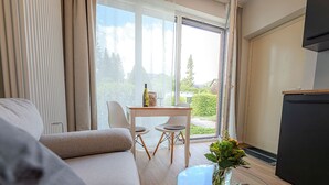 A modern living room with a sofa, a glass coffee table with a vase, a dining table with chairs, and a sliding glass door leading to a balcony. - Studio at the Vineyards (Iphofen)