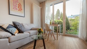 A modern living room with a sofa, a glass coffee table, a dining table with chairs, and a large window with a view of greenery and buildings. - Studio at the Vineyards (Iphofen)