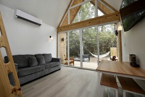A modern interior with a grey sofa, wooden beams, a TV, and a large glass door leading to an outdoor hammock area.