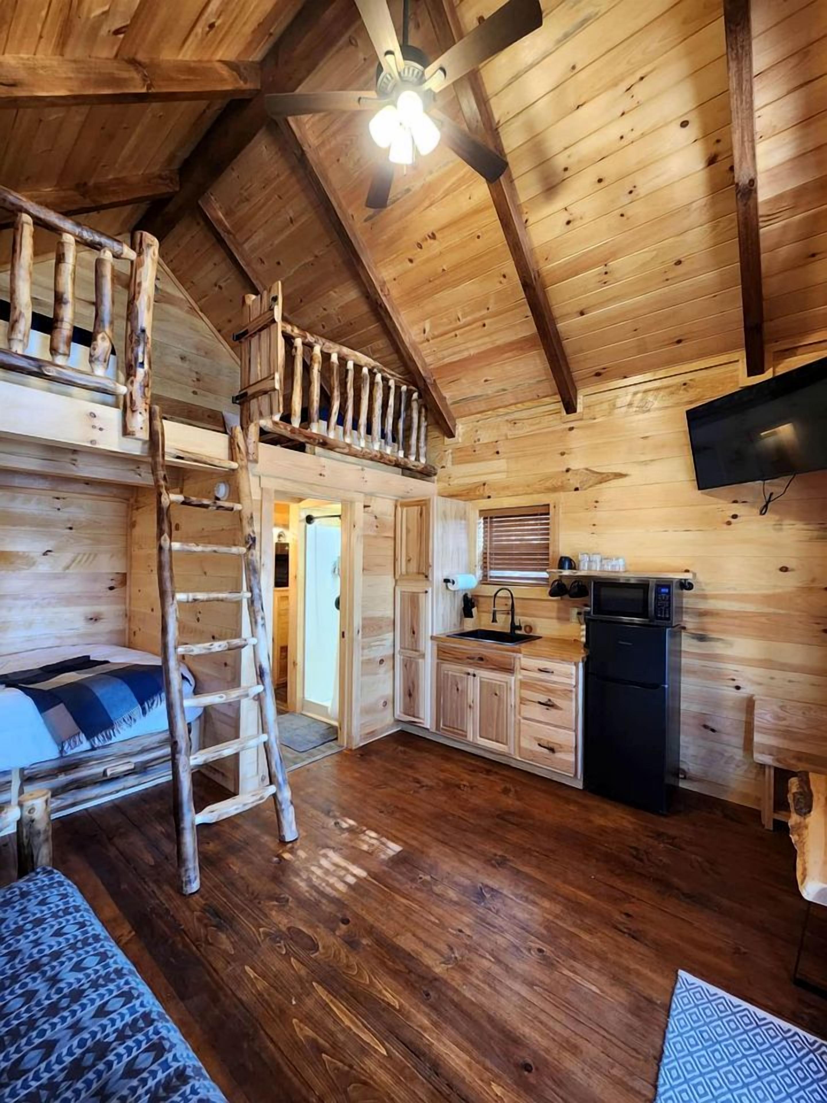 A wooden cabin interior with a loft bedroom, a kitchen area, and a ceiling fan.