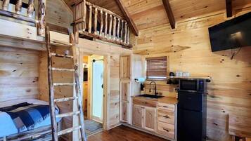 A wooden cabin interior with a loft bedroom, a kitchen area, and a ceiling fan.