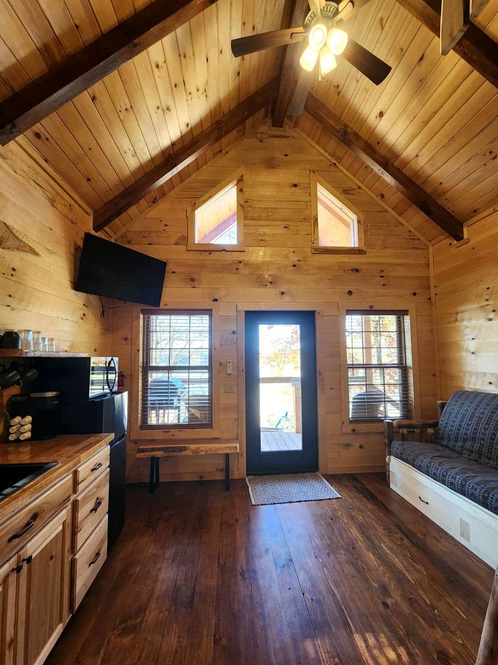 A log cabin interior with a wooden ceiling, a ceiling fan, a television, a sofa, and a kitchen area with a refrigerator and cabinets.