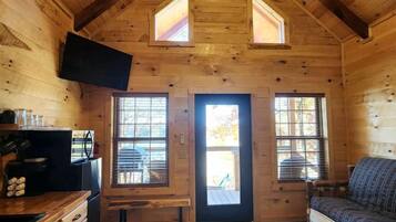 A log cabin interior with a wooden ceiling, a ceiling fan, a television, a sofa, and a kitchen area with a refrigerator and cabinets.