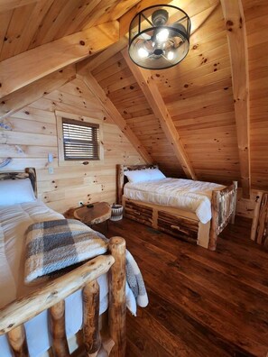 A cozy attic bedroom with two wooden beds, a small table, and a ceiling fan. - Luminous Cabin near Tappan Lake with Hot-Tub, BBQ and Deck in Ohio (Deersville)