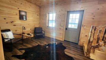 A log cabin interior with a wooden floor, a leather sofa, a chair, a table, a cowhide rug, and a door with windows.