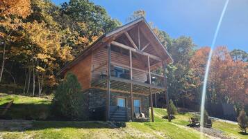 Una cabaña de troncos de dos pisos con un porche cubierto, rodeada de árboles y un cielo despejado.