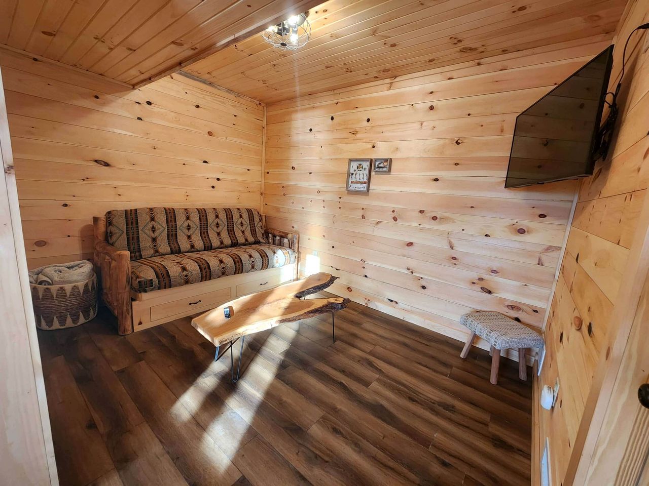 L’intérieur d’un chalet en bois comprend un canapé, une table basse en bois, un petit tabouret et un téléviseur fixé au mur.