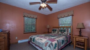 Ein Schlafzimmer mit einem Bett, zwei Fenstern mit Jalousien, einem Deckenventilator, einem Nachttisch mit Lampe und einer Kommode.