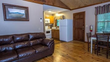 Una sala acogedora con un sofá de cuero marrón, piso de madera, una cocina con armarios de madera, un refrigerador blanco y una mesa de comedor con sillas.