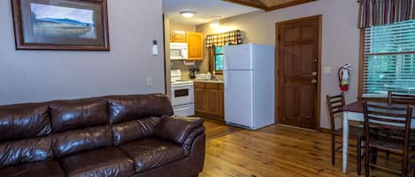 Un salon chaleureux avec un canapé en cuir, un plancher en bois, une cuisine dotée de meubles en bois, un réfrigérateur et un coin repas avec une table et des chaises.