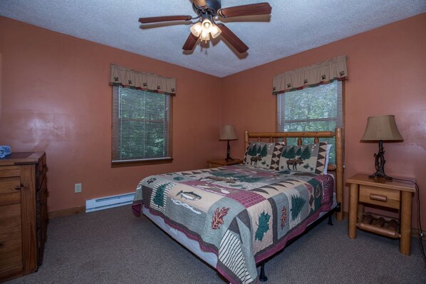 Quarto com cama, duas janelas com persianas, ventilador de teto, mesa de cabeceira com abajur e cômoda.
